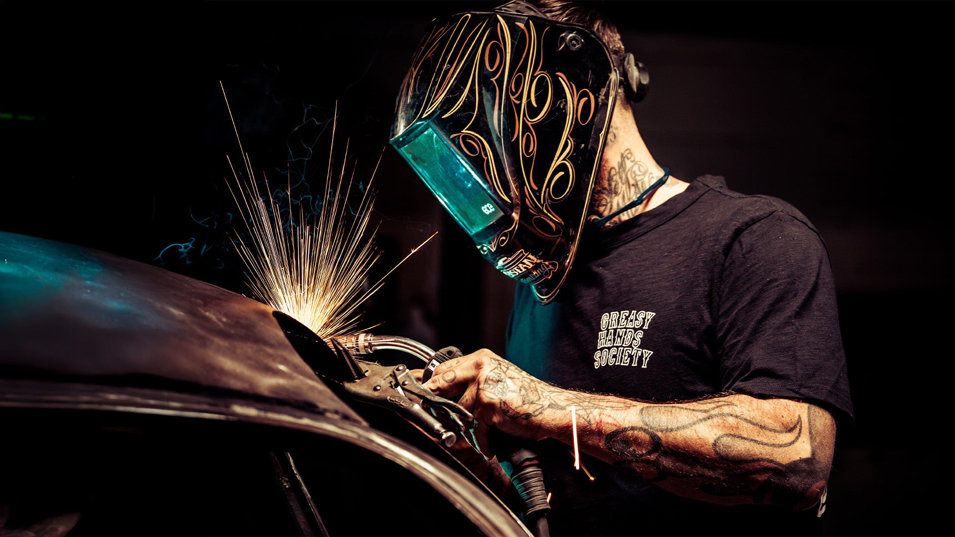 eat dust black graphic tee with greasy hands society on the left chest pocket being worn by a welder, who is welding on his classic car