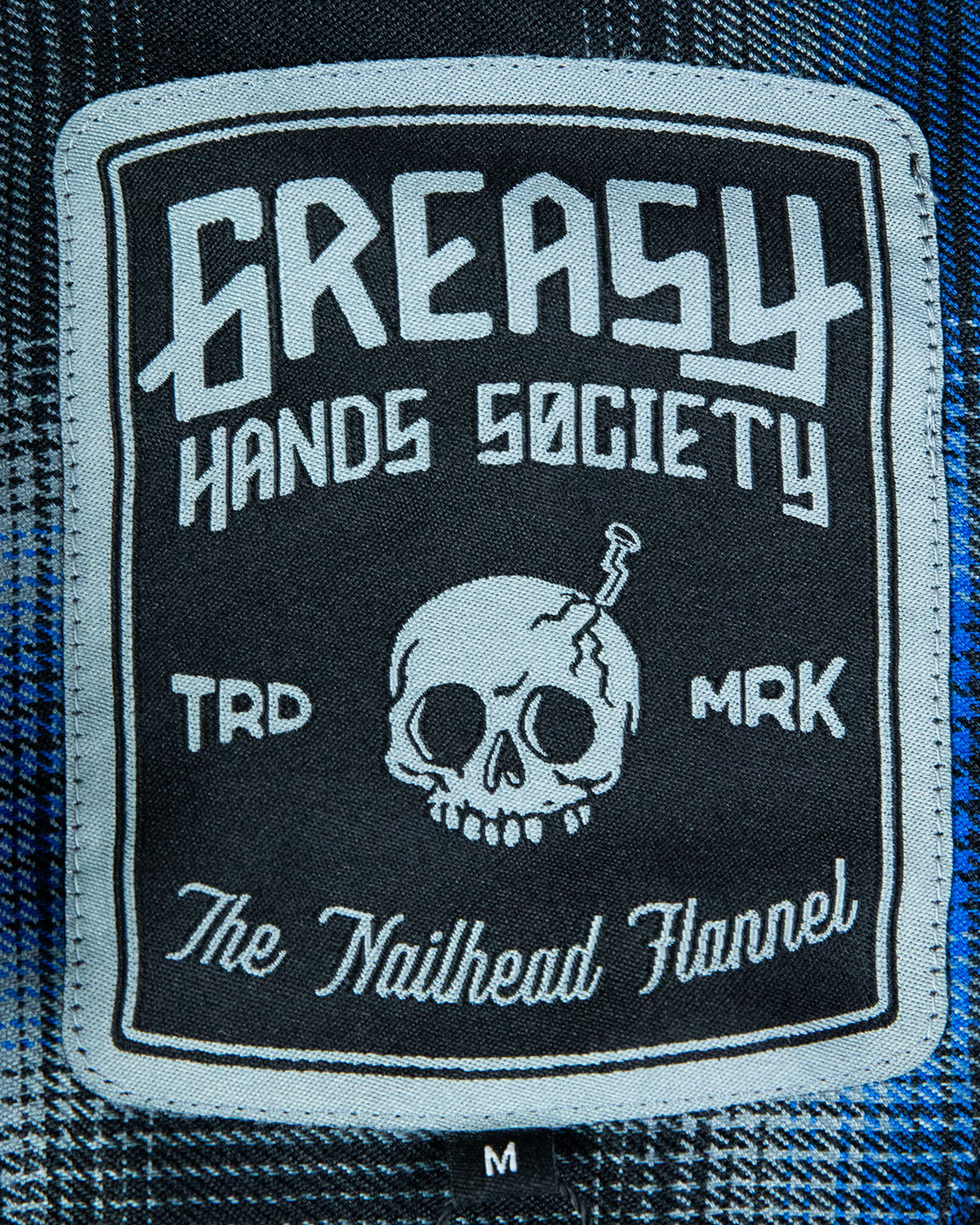 tag Patch on the nailhead blue and black flannel with 'Greasy Hands Society the nailhead flannel' text design with a skull on a flannel fabric background