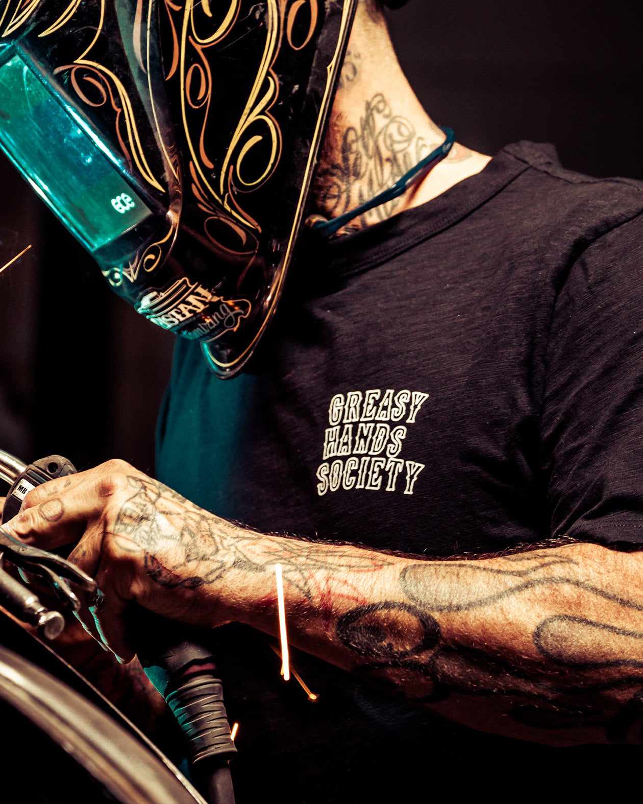 Person wearing a black eat dust graphic tee - t-shirt with 'Greasy Hands Society' text, holding a tool.
