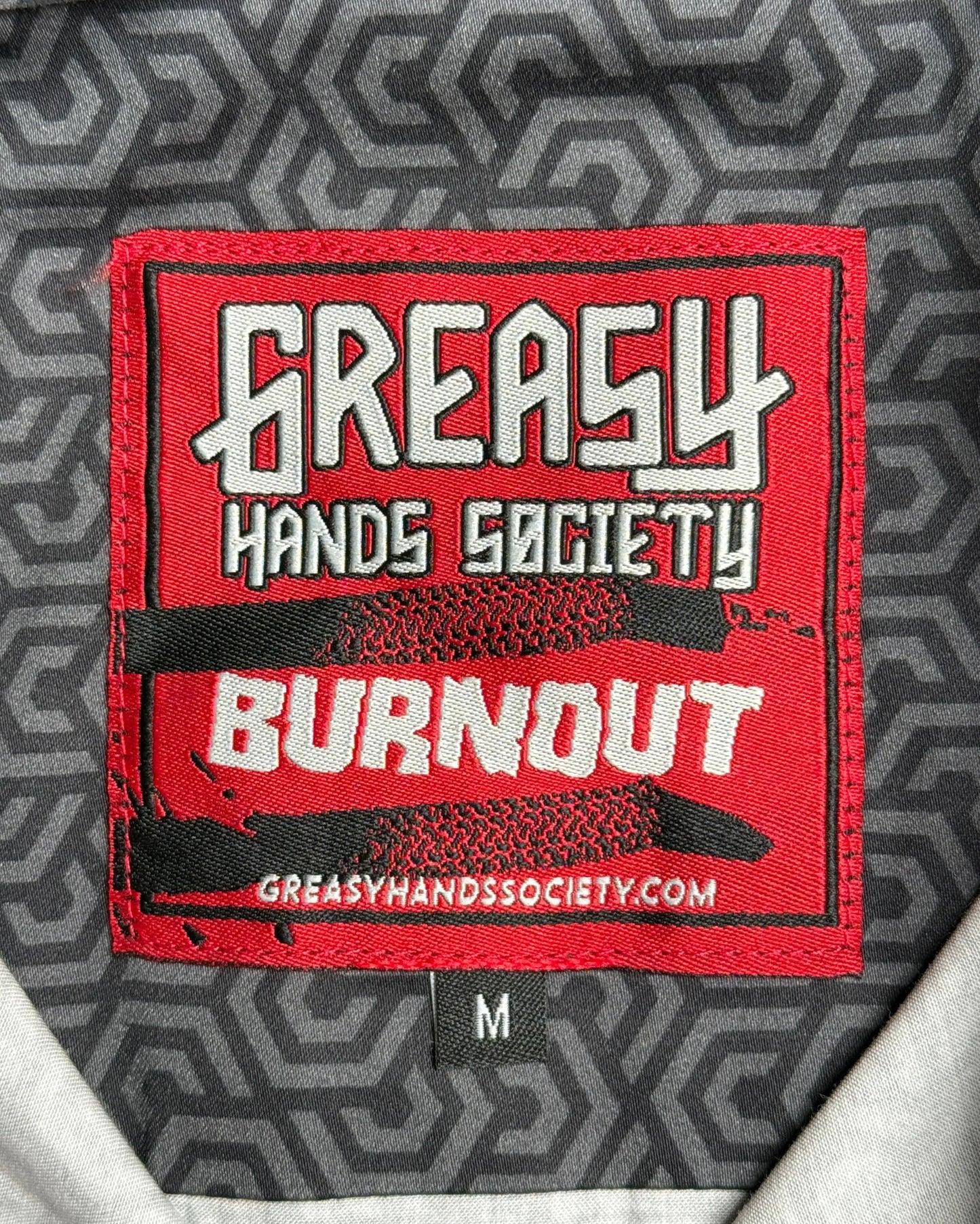 Close-up of a red embroidered patch on a gray patterned garment, reading 'GREASY HANDS SOCIETY' and 'BURNOUT' with a small size tag marked 'M' below - The Burnout Button-Up Shirt by Greasy Hands Society