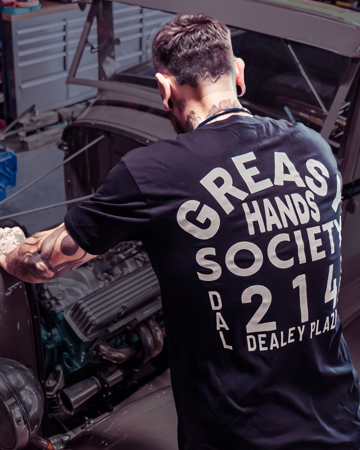 Person wearing a black t-shirt with 'Greas Hands Society' text, standing in a dimly lit room.