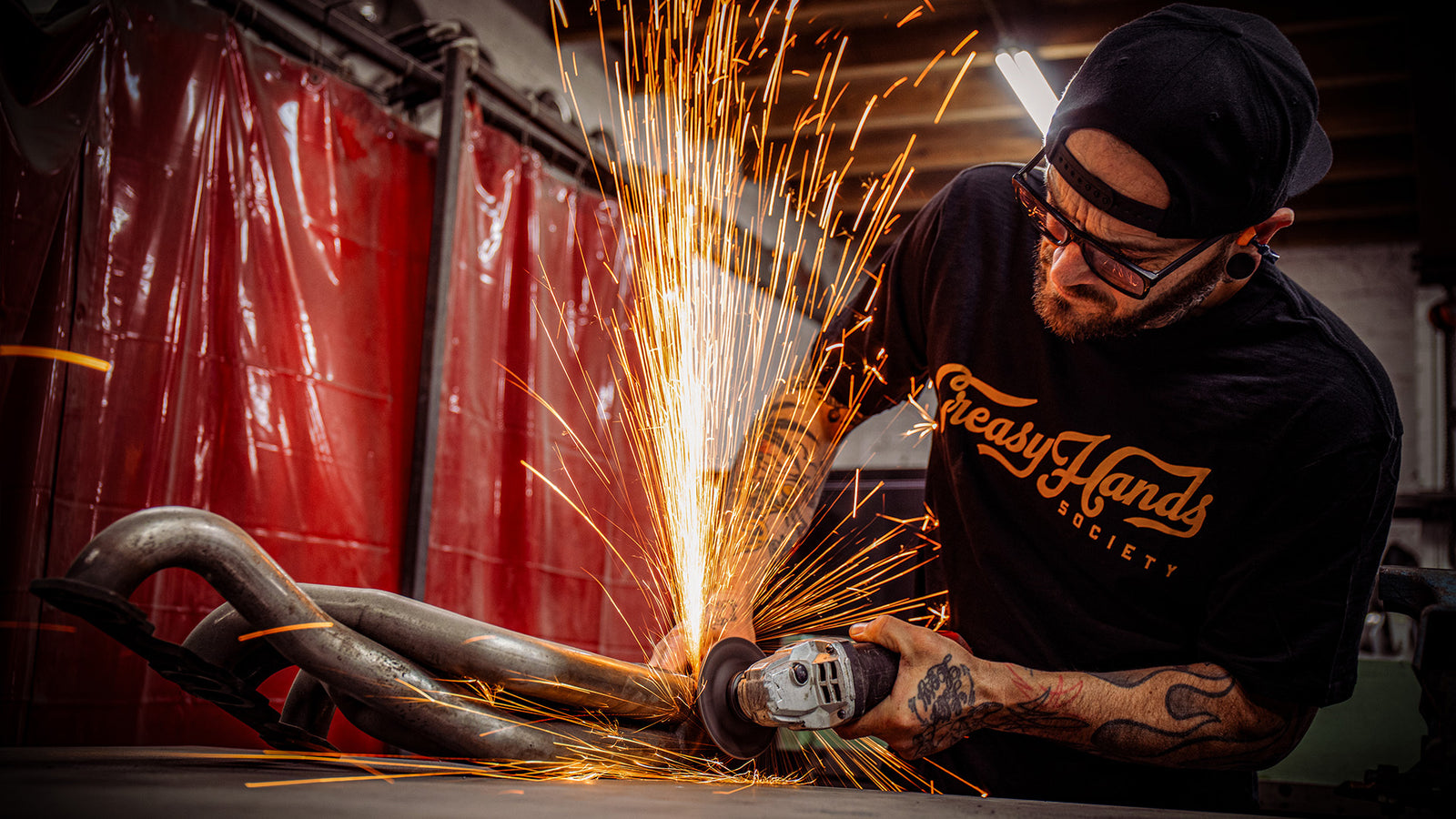 greasy hands society script black t-shirt with gold lettering worn by someone working on car pipes