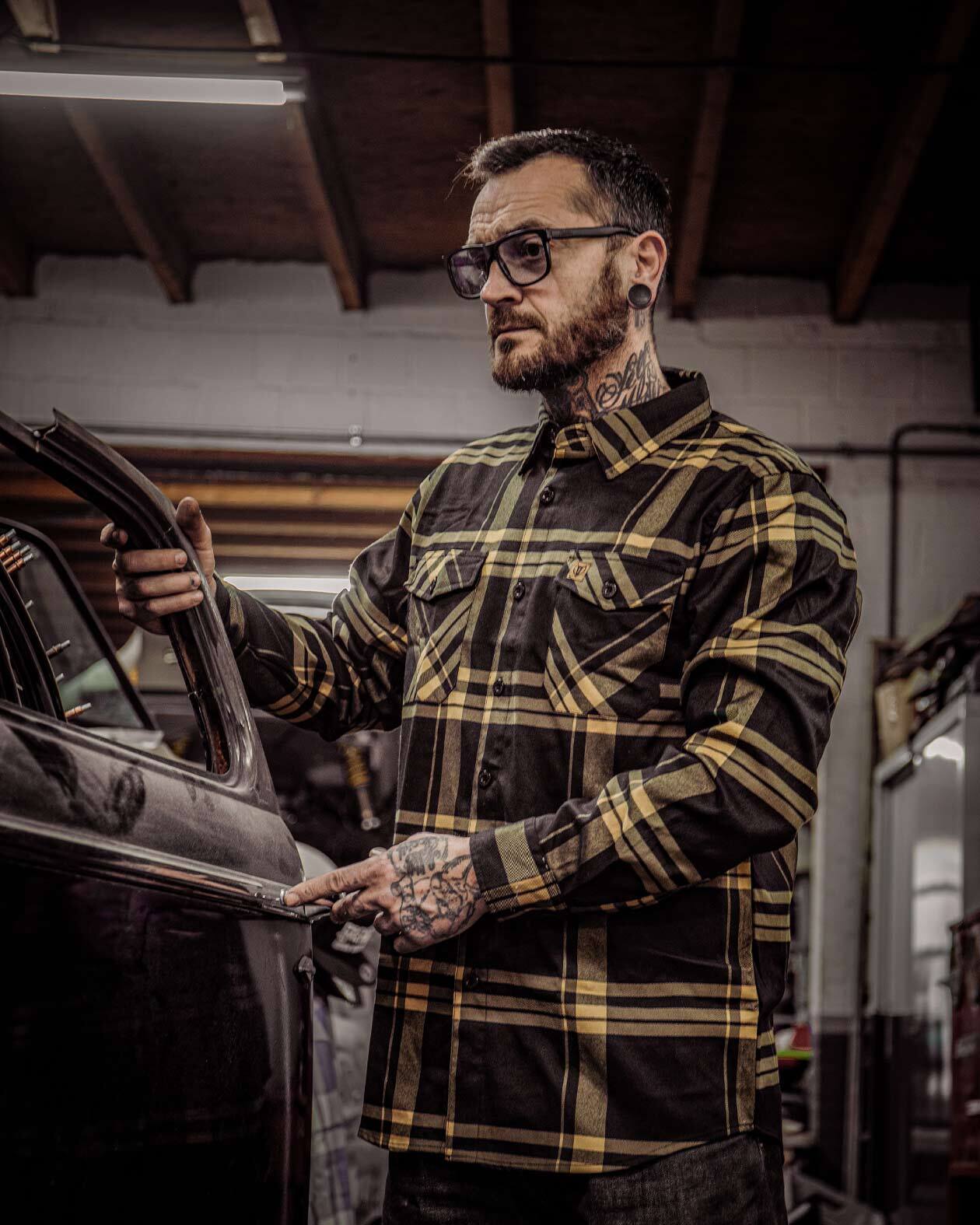 A person with tattoos wearing a black and gold flannel shirt called the two lane flannel with sleeve cuffs and chest pockets, standing near a vehicle inside a garage.