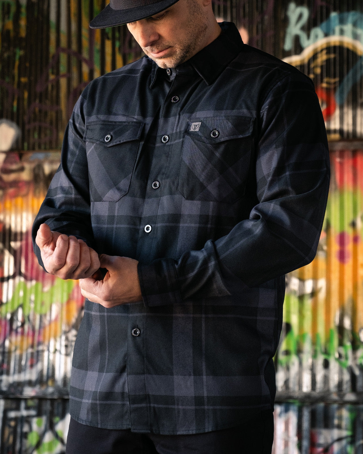 A person wearing a black flannel shirt  called the synister continetal flannel with a geometric pattern, featuring chest pockets and button-up sleeves, standing in front of a colorful background.