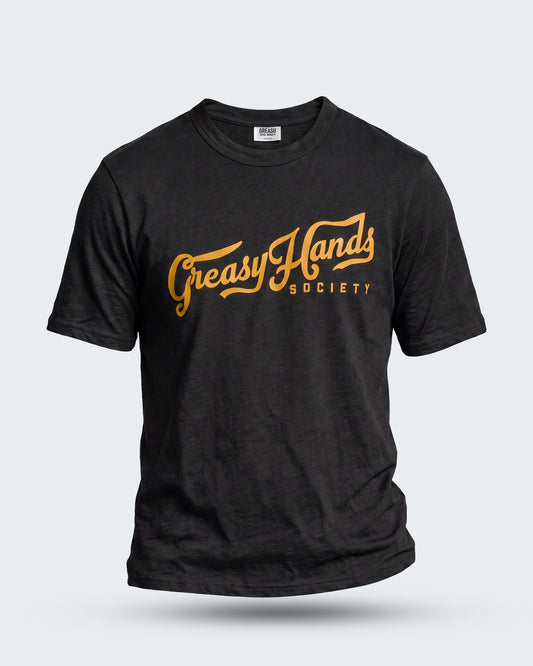 A black short-sleeve t-shirt with a distressed graphic on the chest reading 'Greasy Hands Society' in a golden-yellow cursive font, displayed against a light gray background - Greasy Hands Society Script graphic tee by Greasy Hands Society