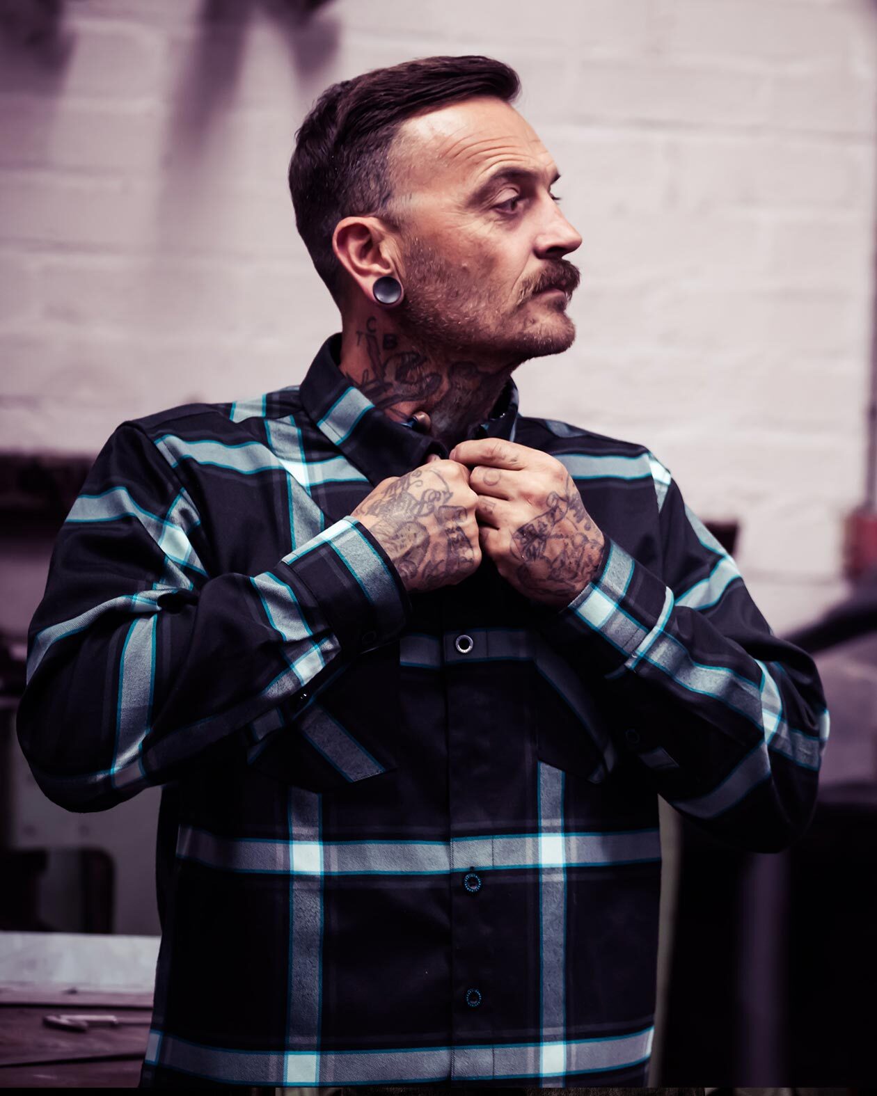 A person wearing a black, blue, and gray plaid flannel shirt called the gravity fed  with hands fixing the collar, standing in a room with white walls.