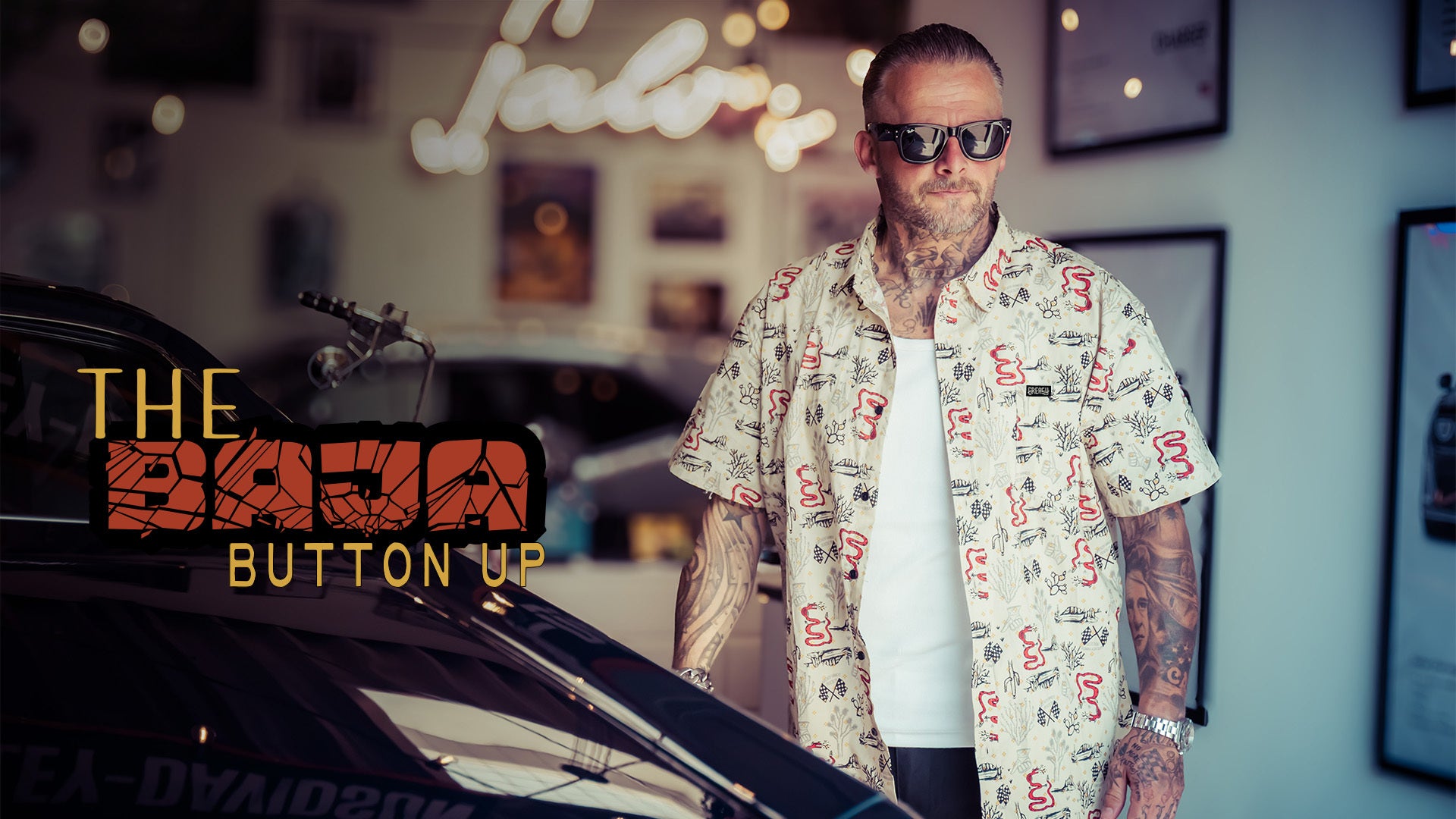 A man wearing sunglasses and a white short-sleeve button-up shirt with a red and black graphic print stands next to a car in a dimly lit indoor setting. Text on the left reads 'THE BAJA BUTTON UP'.