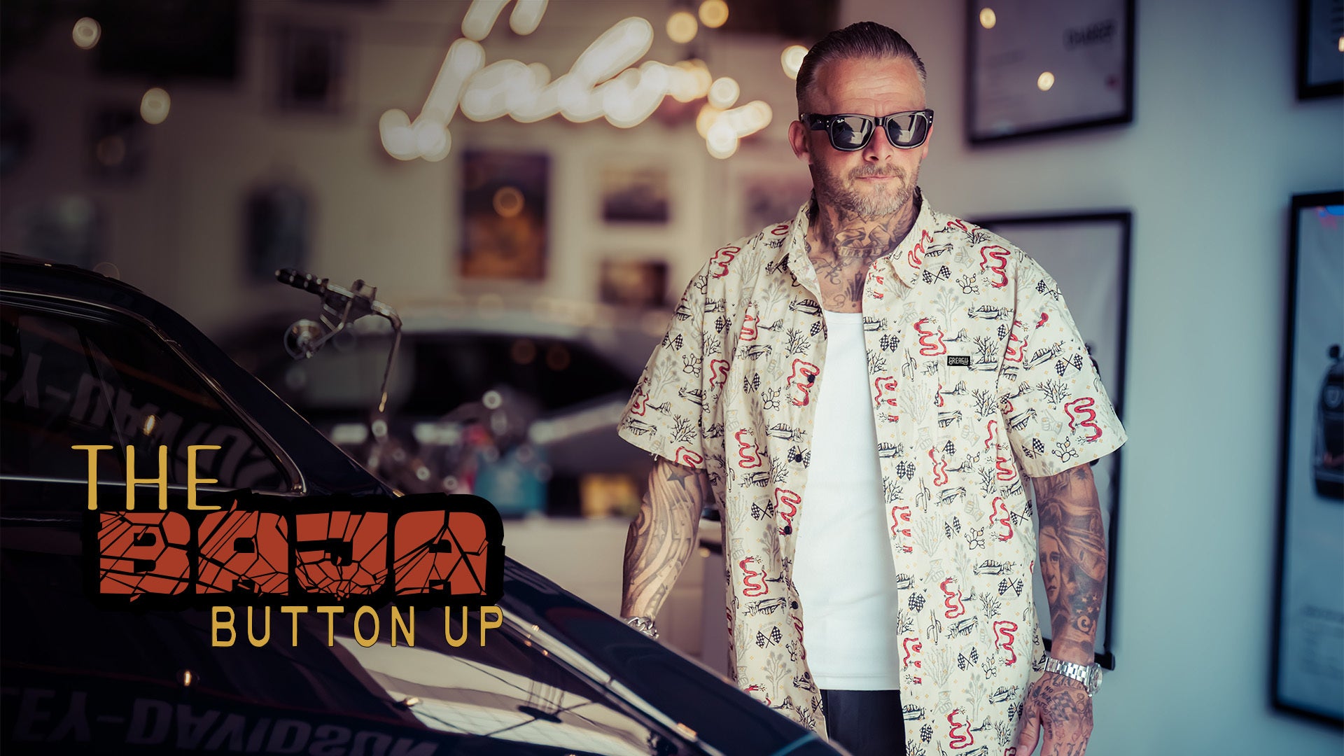 A tattooed man wearing sunglasses and a white short-sleeve button-up shirt with a red and black graphic print stands next to a car in a garage. Text on the left reads 'THE BAJA BUTTON UP'.