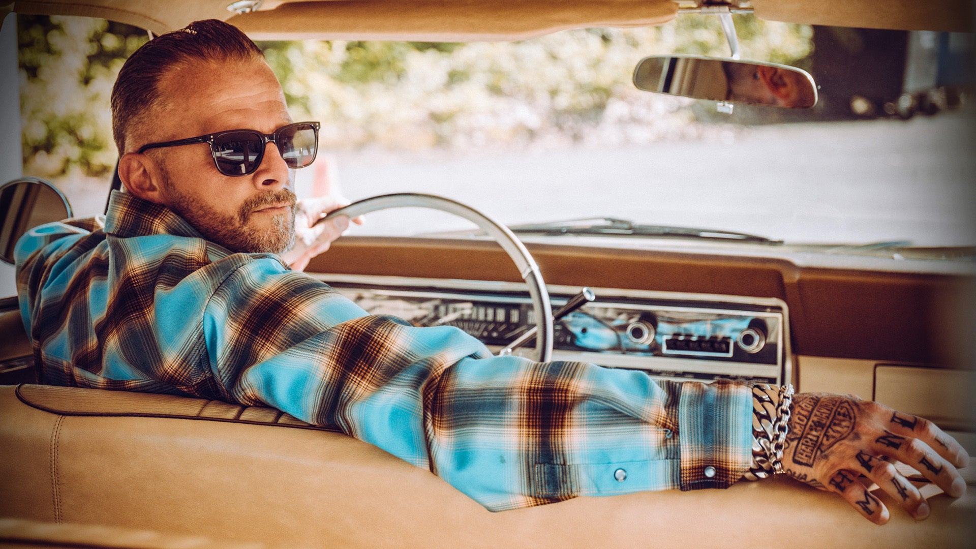 A man wearing black sunglasses and a blue, brown, and black plaid flannel shirt leans back in a vintage car interior, holding a cigarette.
