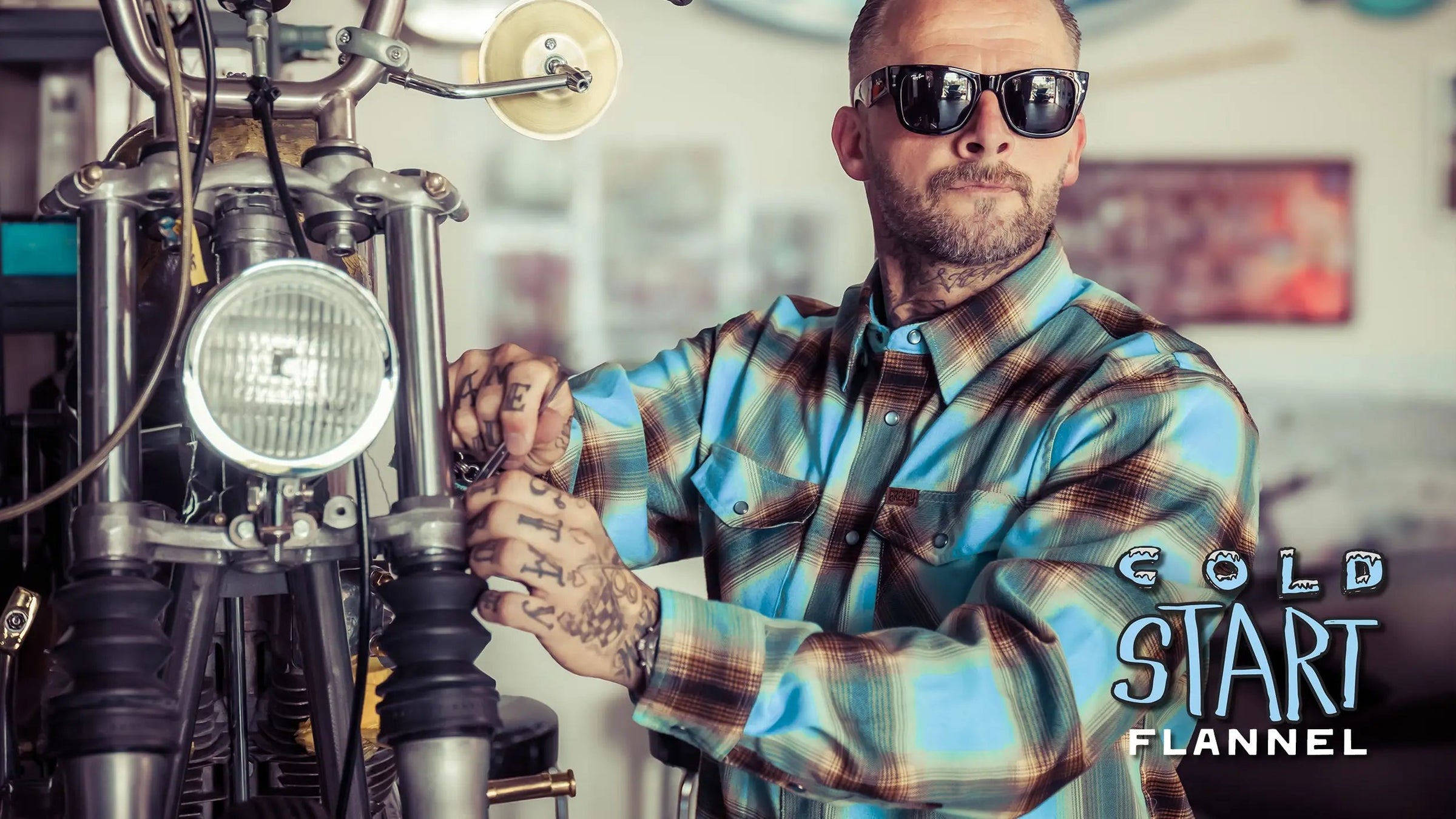 A man wearing a blue, brown, and beige plaid button-down shirt and black sunglasses stands next to a motorcycle in a workshop setting. The text 'COLD START FLANNEL' is visible in the bottom right corner - The Cold Start Flannel by Greasy Hands Society