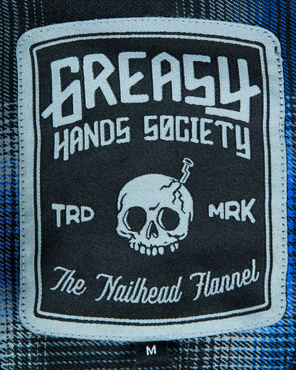 tag Patch on the nailhead blue and black flannel with 'Greasy Hands Society the nailhead flannel' text design with a skull on a flannel fabric background