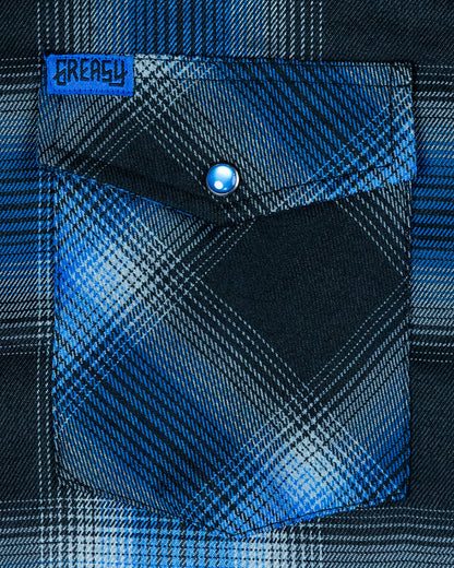 Close-up of a blue and black plaid flannel shirt pocket with a button, featuring the brand 'Greasy' - called the nailhead flannel - The Nailhead Flannel by Greasy Hands Society