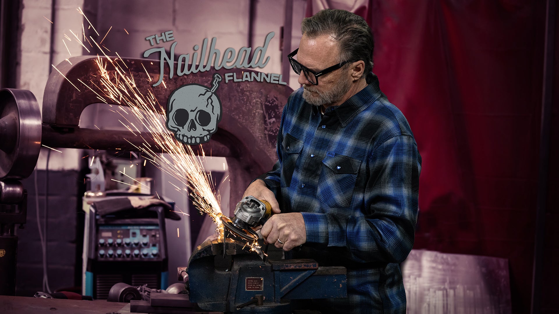 Man using a grinder with sparks in a workshop setting, wearing protective glasses. wearing a blue and black plaid flannel with "The Nailhead Flannel" branding.