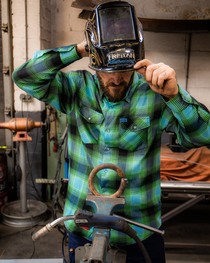 Person wearing a green and blue plaid flannel shirt and welding helmet in a workshop setting - Sucker Punch Flannel by Greasy Hands Society