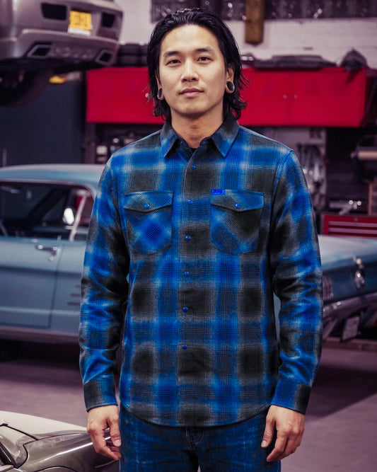 Man wearing a blue black and gray plaid flannel shirt standing in a garage with cars in the background - The Nailhead Flannel by Greasy Hands Society