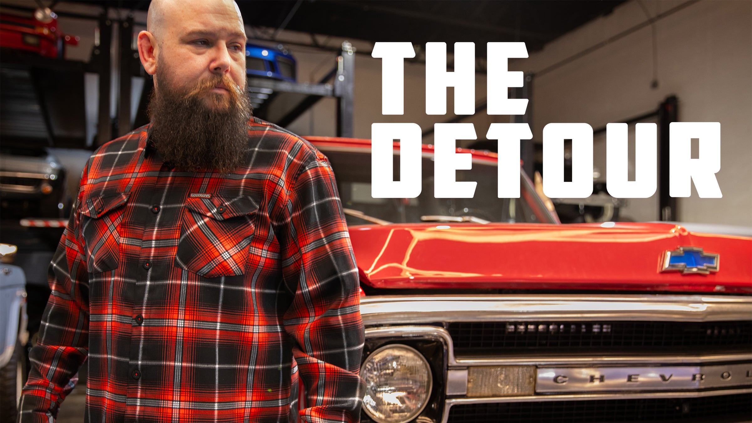 Man with a beard standing next to a red Chevrolet car in a garage with 'The Detour' text.