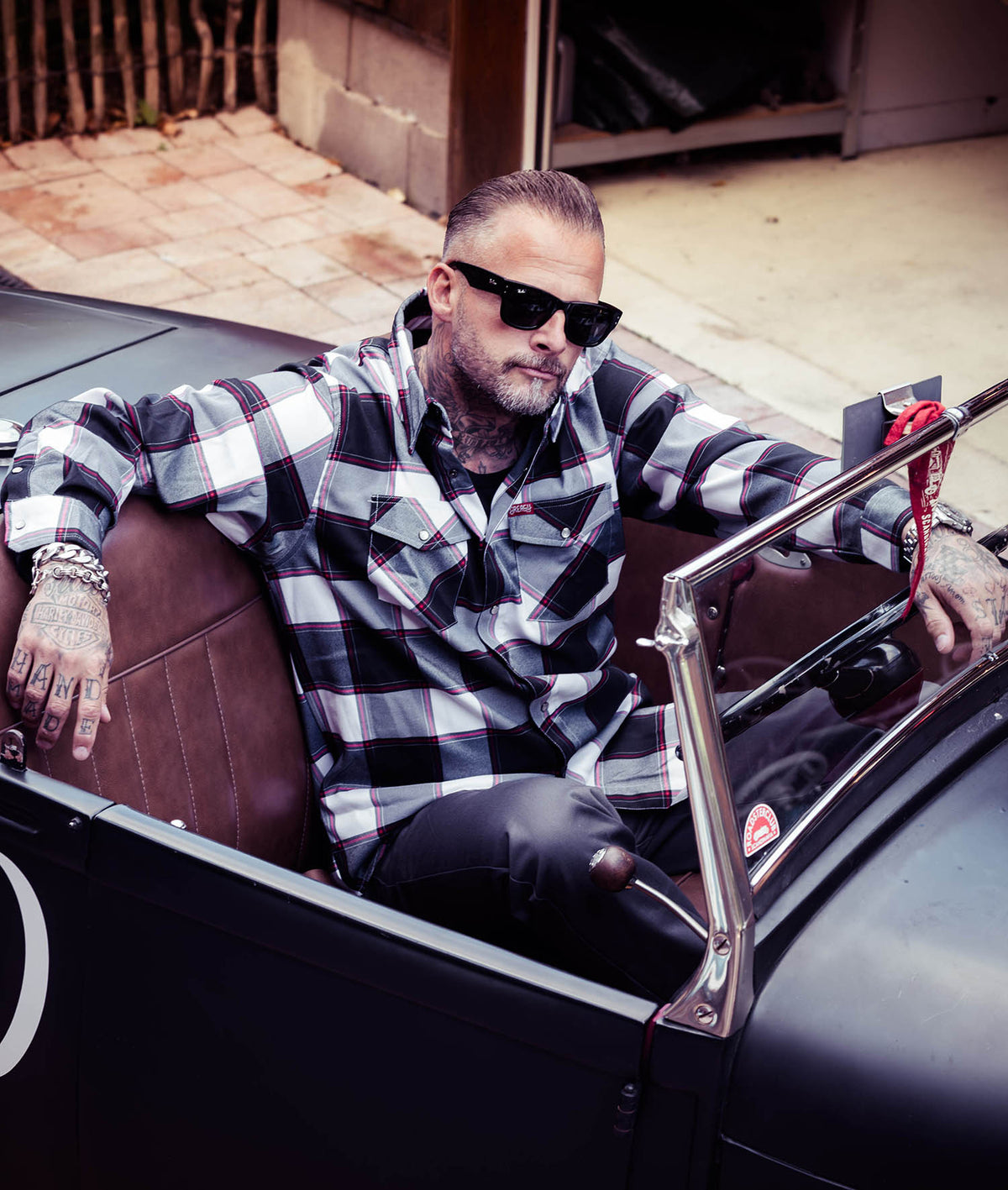 A man wearing a black and white buffalo plaid button-down flannel shirt and black sunglasses sits in a vintage black convertible car with brown leather seats - The Suicide Machine Flannel by Greasy Hands Society
