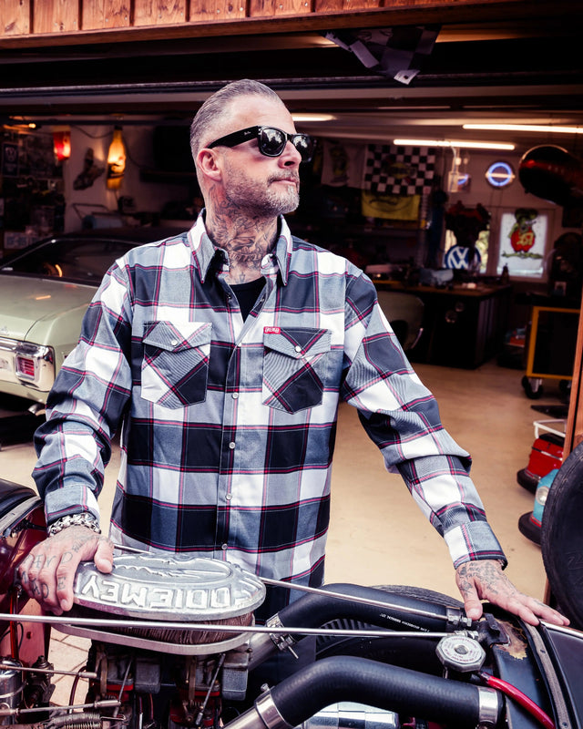 A man with tattoos and sunglasses stands in a garage wearing a black, white, and red plaid button-down flannel shirt, leaning on a motorcycle engine - Suicide Machine Flannel by Greasy Hands Society