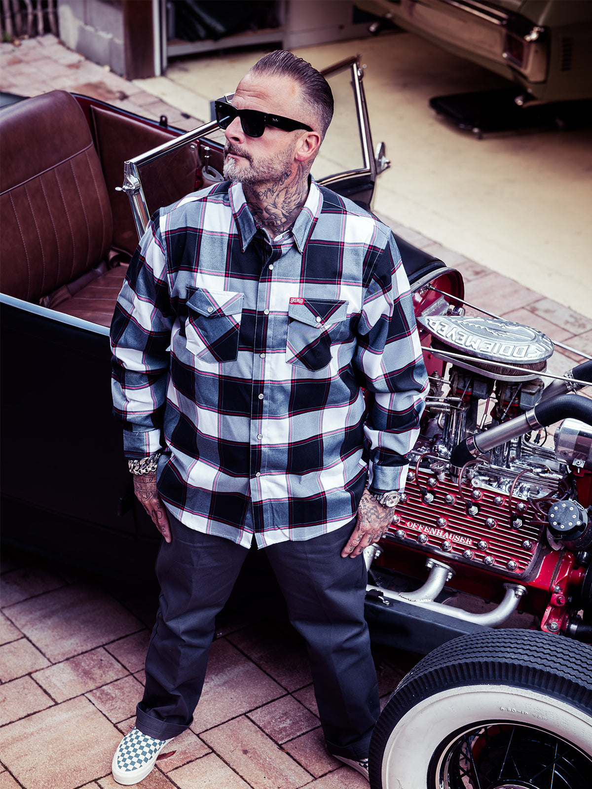 A man wearing a black and white buffalo plaid button-down flannel shirt and black sunglasses standing outside of a vintage black convertible car with brown leather seats - The Suicide Machine Flannel by Greasy Hands Society