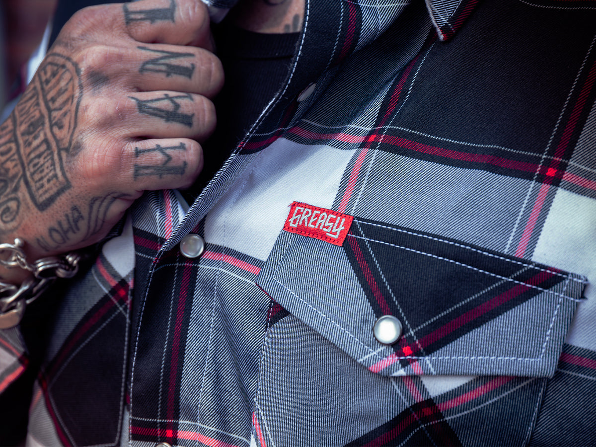 Close-up of a person wearing a gray, black, and red plaid flannel shirt, with a red embroidered 'GREASY' patch on the chest pocket - The Suicide Machine Flannel by Greasy Hands Society