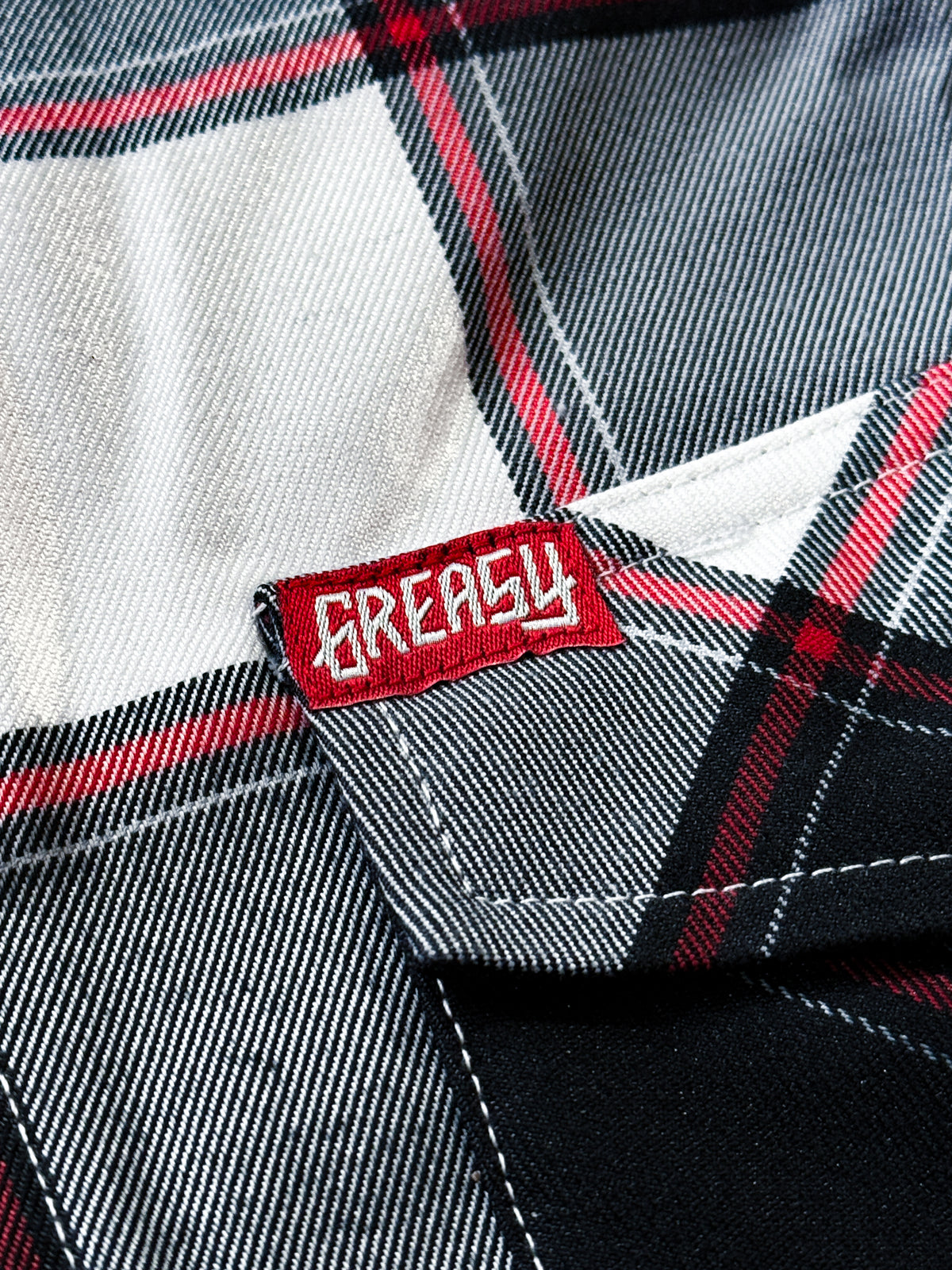 Close-up of a black, white and red plaid button-up flannel shirt fabric with a red embroidered patch label reading 'GREASY' in white text - Suicide Machine Flannel by Greasy Hands Society