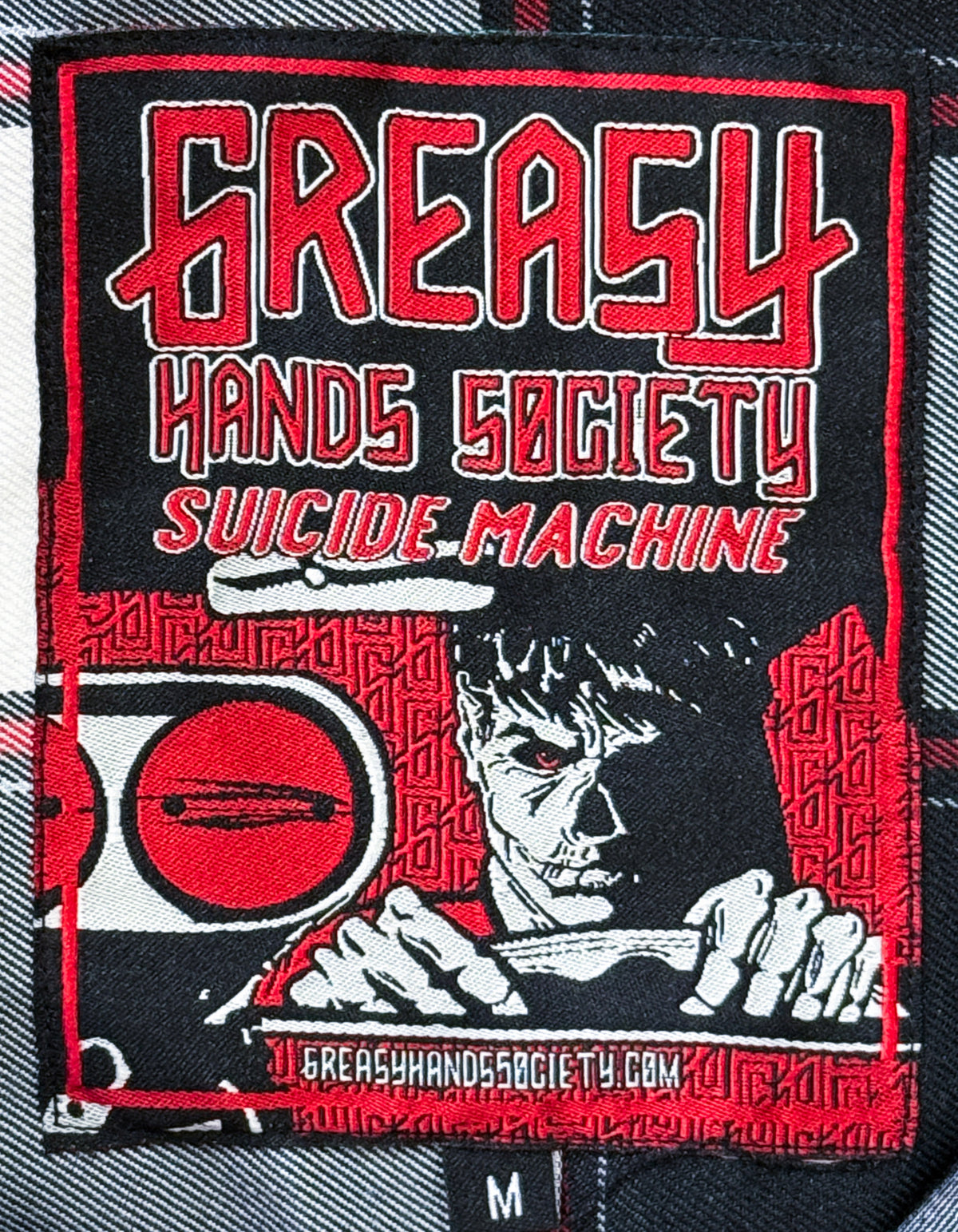 A close-up of a black and red embroidered tag for a Greasy Hands Society flannel, featuring the text 'GREASY HANDS SOCIETY' and 'SUICIDE MACHINE' with a graphic illustration of a face and hands holding a knife, set against a plaid fabric background - Suicide Machine Flannel by Greasy Hands Society