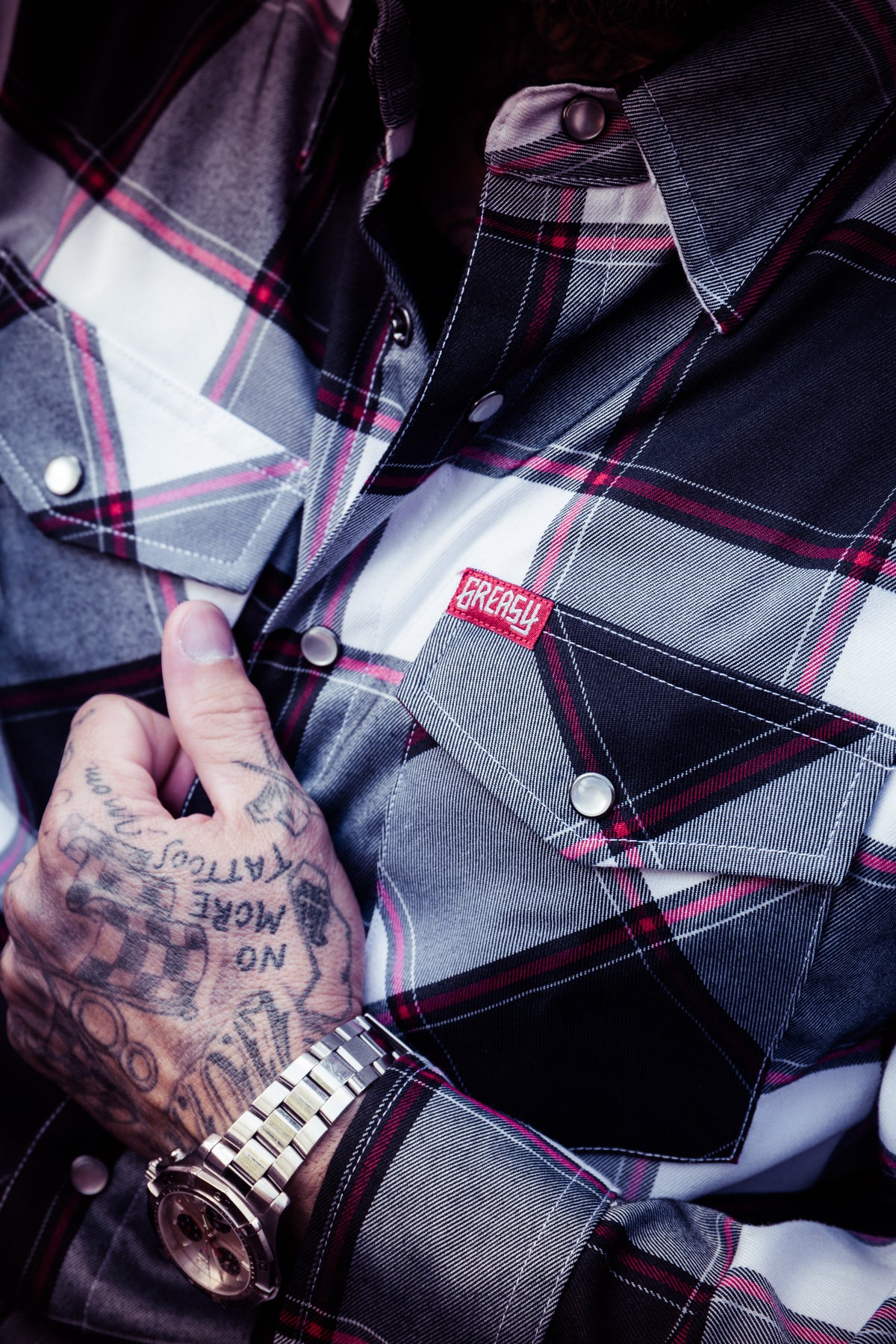 A close-up of a person wearing a plaid button-up flannel shirt with a red and white embroidered chest patch. The shirt has a gray, black, and white tartan pattern with red lines, and the person's tattooed hand is resting near the pocket - The Suicide Machine Flannel by Greasy Hands Society