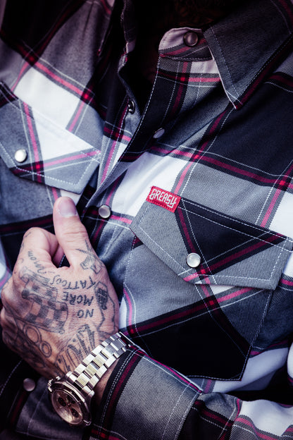 A close-up of a person wearing a plaid button-up flannel shirt with a red and white embroidered chest patch. The shirt has a gray, black, and white tartan pattern with red lines, and the person's tattooed hand is resting near the pocket - The Suicide Machine Flannel by Greasy Hands Society