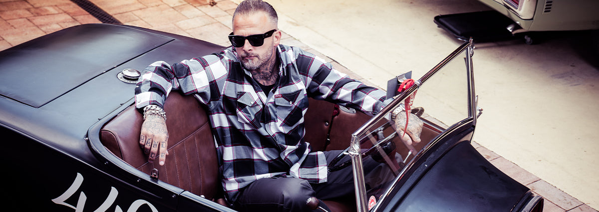 A man wearing a black and white plaid flannel shirt and sunglasses sits in the open cockpit of a black vintage convertible car with brown leather seats - The Suicide Machine Flannel by Greasy Hands Society