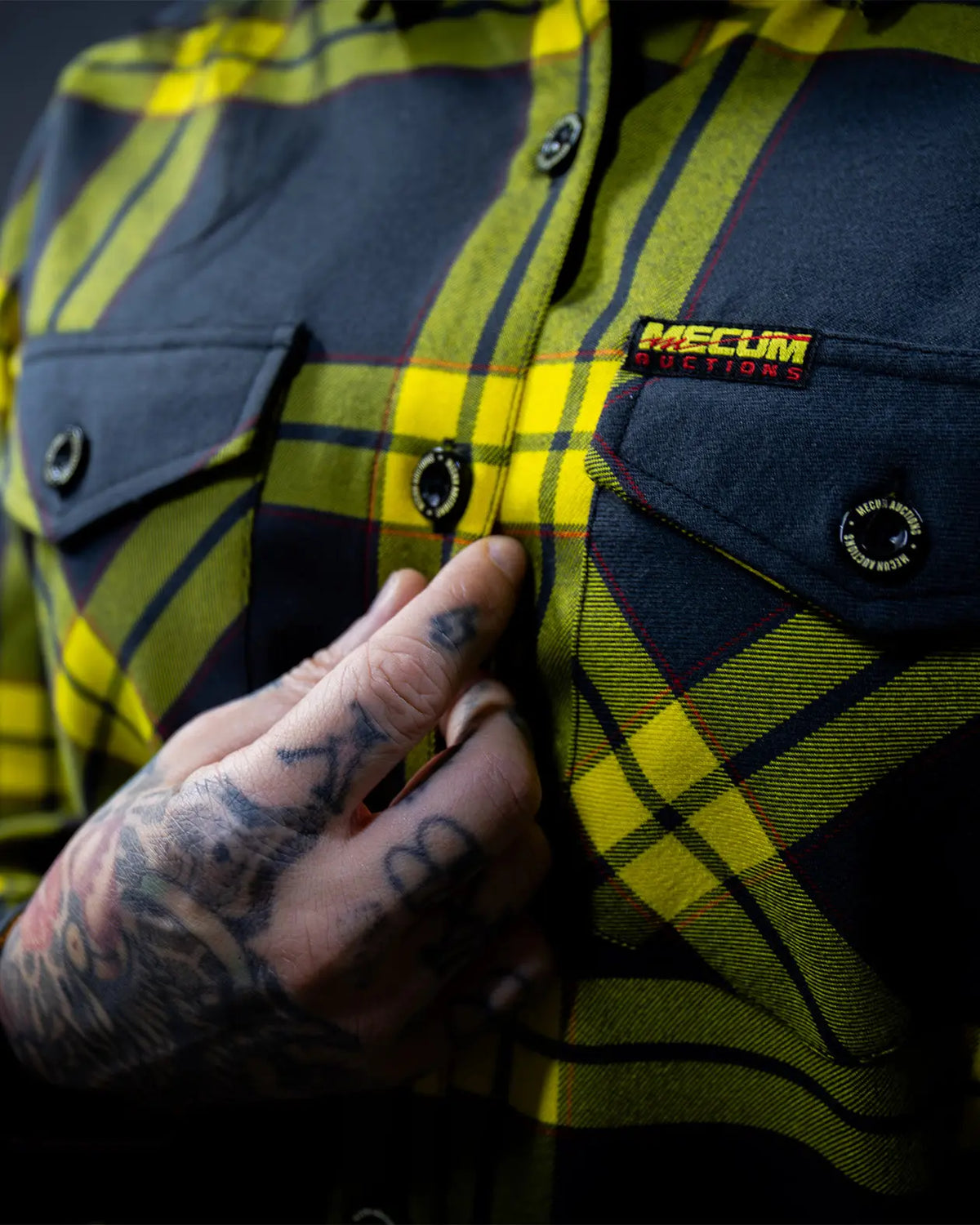 Close-up of a person wearing a dark gray and yellow plaid flannel button-up shirt, with a tattooed hand adjusting the collar. A 'MECUM AUCTIONS' brand patch is visible on the chest pocket - The Main Attraction Flannel - Mecum Auctions x Greasy Hands Society Collab Flannel