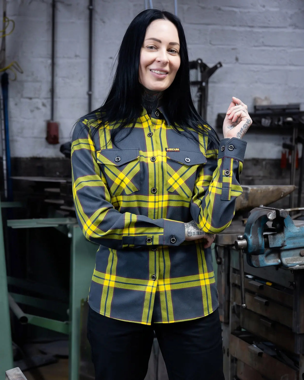 A woman with long black hair stands in a workshop wearing a dark gray and yellow plaid button-up flannel shirt with black pants. The shirt features a small yellow 'MECUM AUCTIONS' logo on the left chest pocket  - The Main Attraction Flannel - Mecum Auctions x Greasy Hands Society Collab Flannel