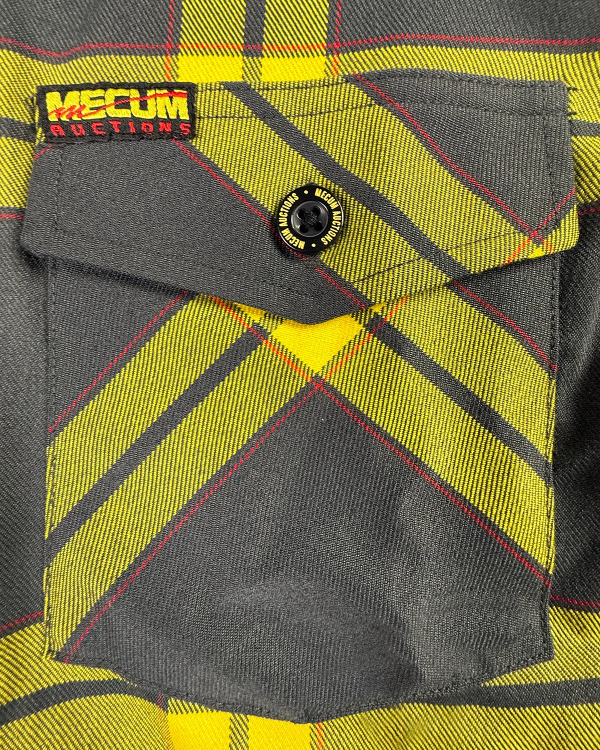 Close-up of a plaid flannel shirt pocket with a yellow, gray, and red pattern, featuring a flap pocket and a black button. A 'MECUM AUCTIONS' brand label is visible on the upper left - The Main Attraction Flannel - Mecum Auctions x Greasy Hands Society Collab Flannel