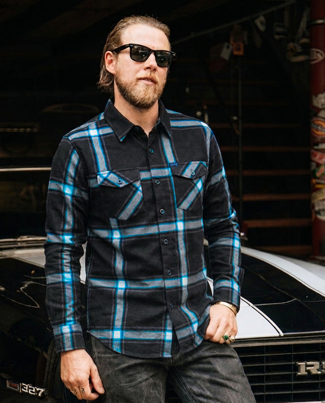 Man wearing a plaid black and blue flannel shirt in front of a car - The Gravity Fed Flannel by Greasy Hands Society