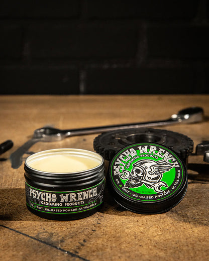 an open containers of Psycho Wrench grooming oil-based pomade on a wooden surface with tools in the background
