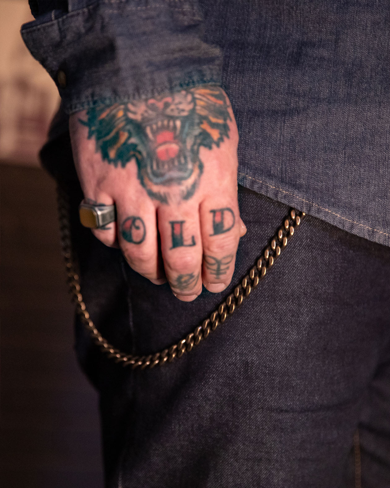 Hand with tattoos holding a chain for a wallet, wearing a gold ring, against a dark denim background.