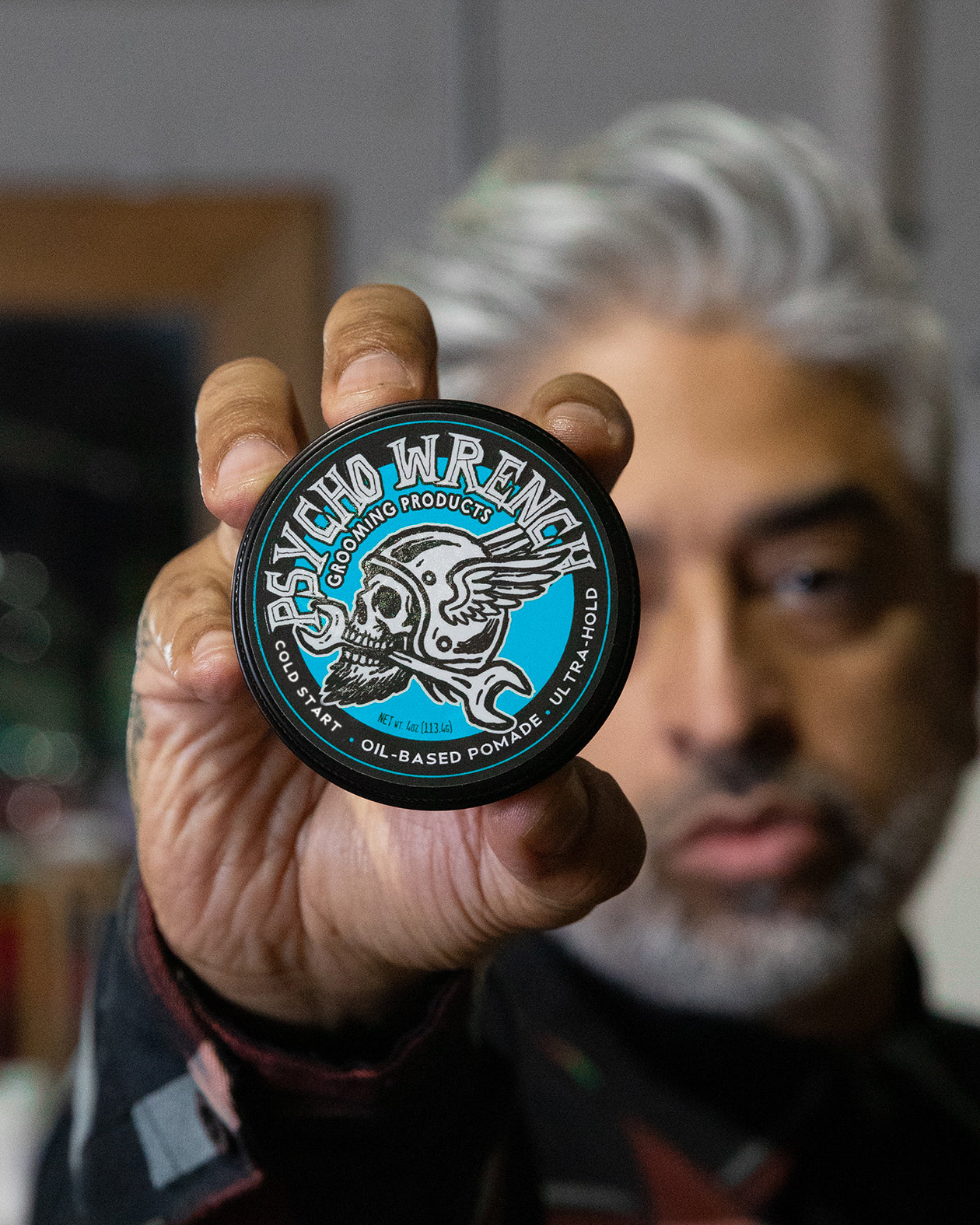 Person holding a container of 'Psycho Wrench' pomade with a blurred background - Psycho Wrench Cold Start Pomade by Greasy Hands Society