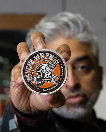 Person holding a product container with a brand logo against a blurred background - Psycho Wrench Tap & Die Clay Pomade by Greasy Hands Society