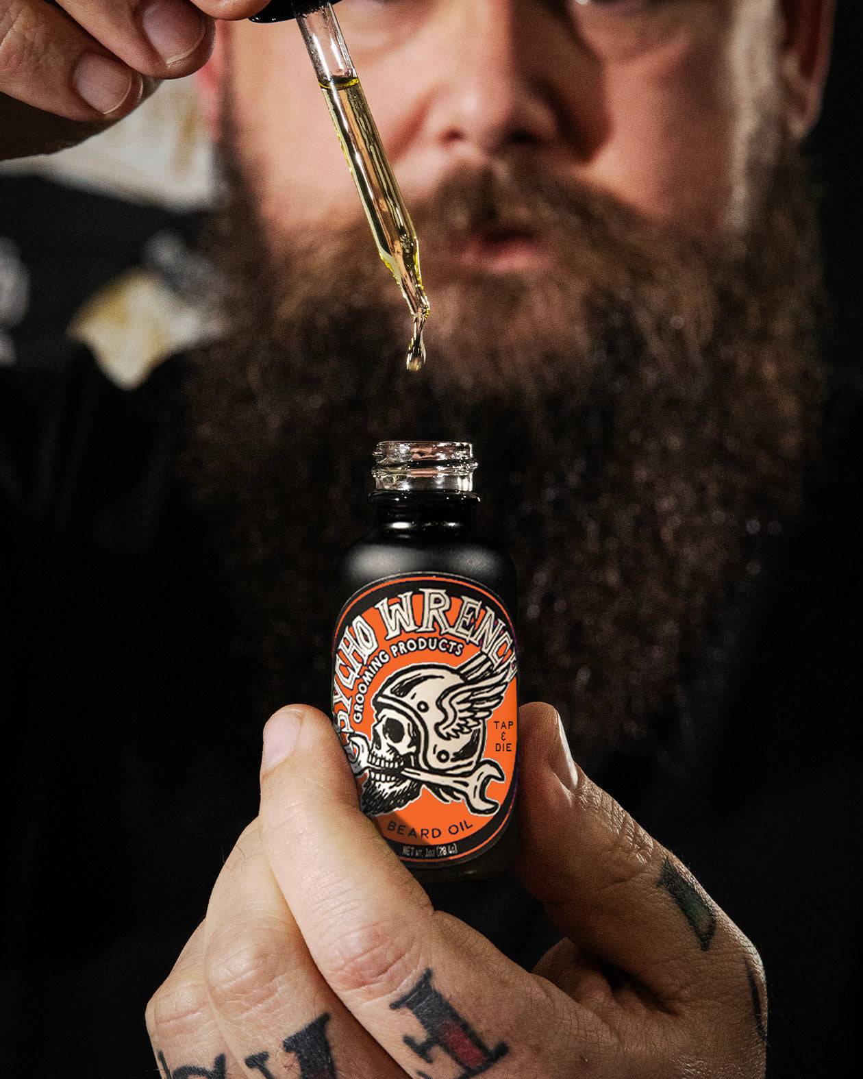 Man holding a bottle of 'Psycho Wrench Grooming Product' beard oil with a dropper above it.