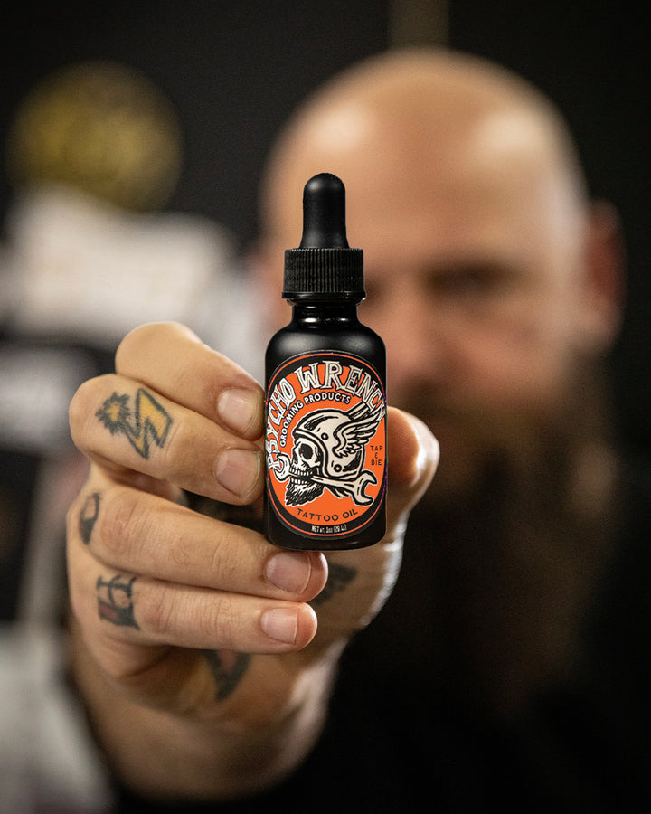 Person holding a bottle of 'Psycho Wrench' beard oil with a blurred background - Psycho Wrench Tap & Die Tattoo Oil by Greasy Hands Society