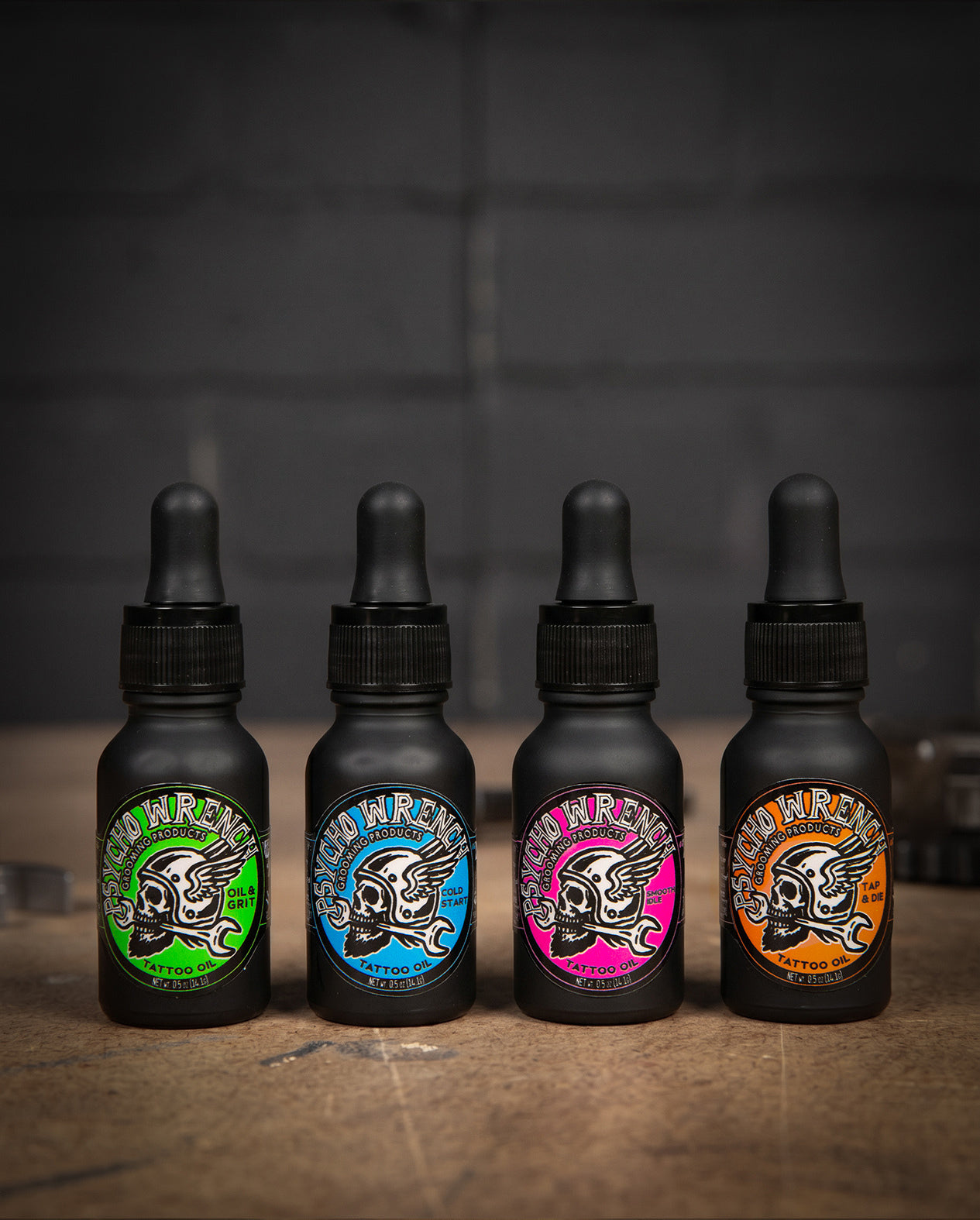 Four black bottles with colorful labels on a wooden surface and dark background - Tattoo Oil variety 4-pack - Oil & Grit, Cold Start, Smooth Idle, and Tap & Die - Tattoo Oil Variety Pack by Greasy Hands Society