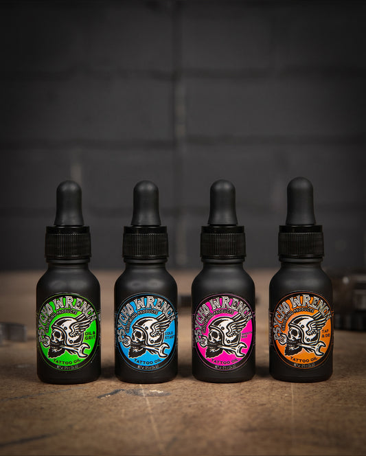 Four black bottles with colorful labels on a wooden surface and dark background - Tattoo Oil variety 4-pack - Oil & Grit, Cold Start, Smooth Idle, and Tap & Die - Tattoo Oil Variety Pack by Greasy Hands Society