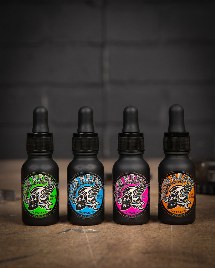 Four black bottles with colorful labels on a wooden surface and dark background - Tattoo Oil variety 4-pack - Oil & Grit, Cold Start, Smooth Idle, and Tap & Die - Tattoo Oil Variety Pack by Greasy Hands Society