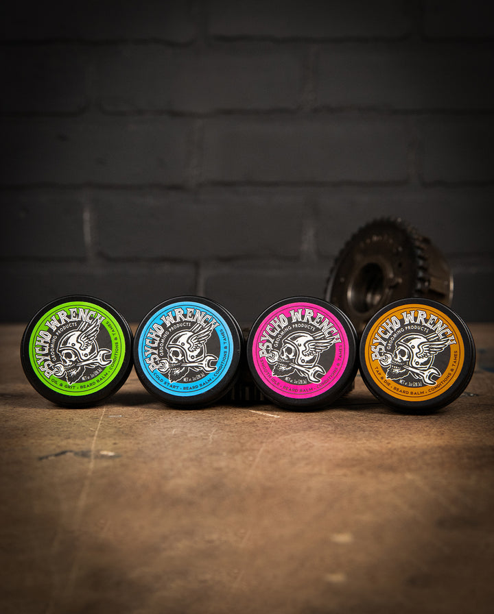 Four circular products with colorful labels on a wooden surface and dark brick wall background - Beard Balm Variety 4-pack by greasy hands society