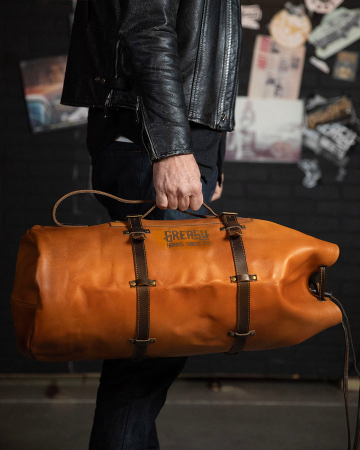 Person holding a brown leather duffel bag for men with 'Greasy Hands Society' branding against a dark background - The Freeloader Duffel Bag from greasy hands society