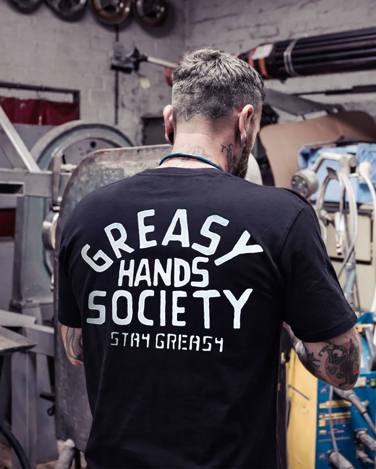 Person wearing a black t-shirt with 'Greasy Hands Society Stay Greasy' text in a workshop setting