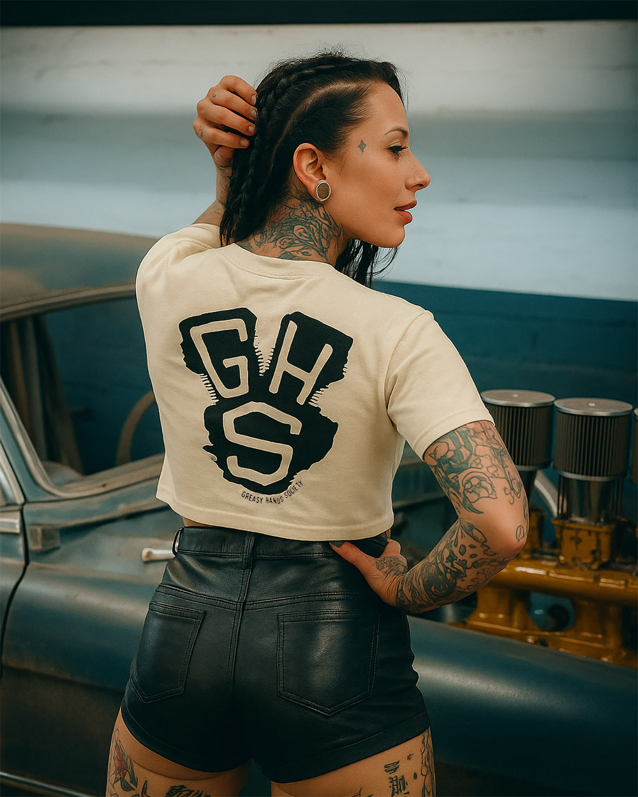 woman wearing a white t-shirt with 'GHS' on the back with an engine sillhouetted in the back and leather shorts, standing next to a vintage car.