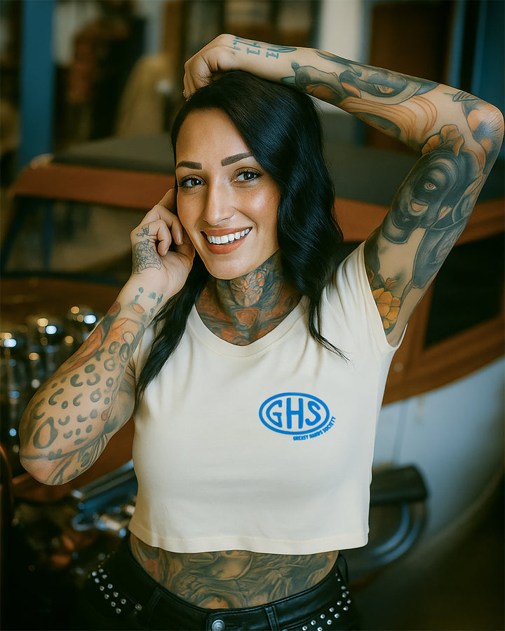 Woman in a beige crop top with "GHS" on the left chest in an industrial setting -Buckle Up Crop Top by Greasy Hands Society