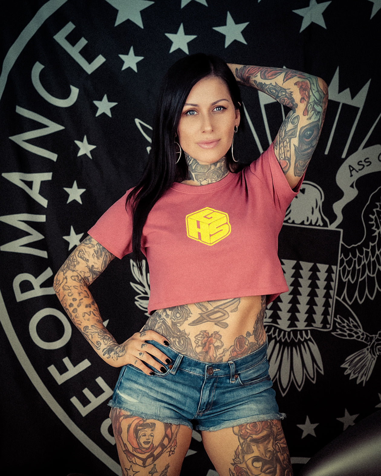 Woman with tattoos wearing a peach crop top with GHS small block design and denim shorts in front of a black background with white text and designs.