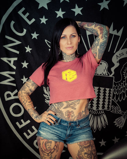 Woman with tattoos wearing a peach crop top with GHS small block design and denim shorts in front of a black background with white text and designs.