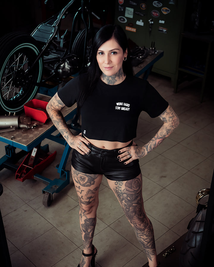 Woman wearing a black crop top t-shirt with 'Work Hard, Stay Greasy' text, standing in front of colorful vehicles.